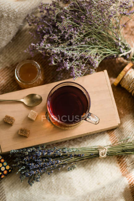 Bunches of dried herbs and jar of fresh honey on blanket near board with cup of hot tea and cubes of sugar. — Stock Photo