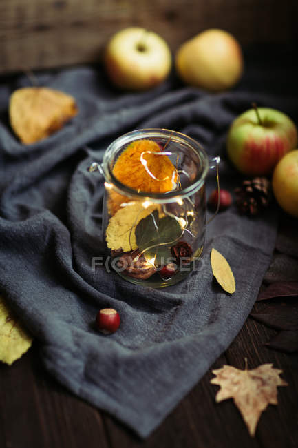 Piccole noci e foglie secche autunnali poste in vaso di vetro con luci fiabesche su un pezzo di tessuto vicino alle mele fresche . — Foto stock