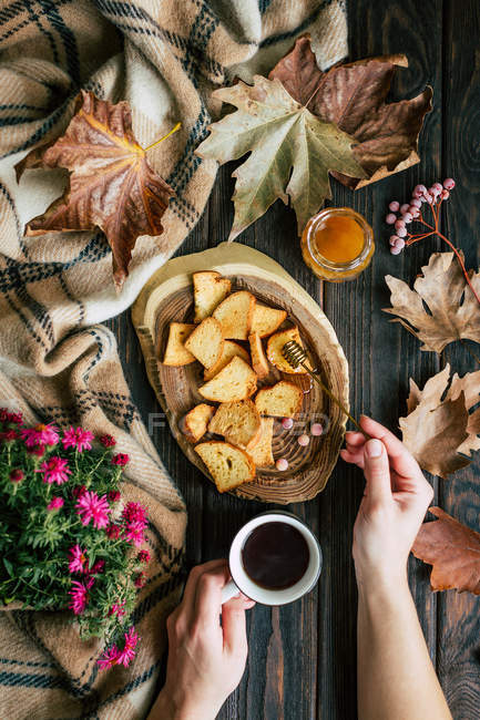 De cima mãos de mulher segurando xícara de chá quente e espalhando mel fresco em croutons crocantes perto de folhas de outono . — Fotografia de Stock