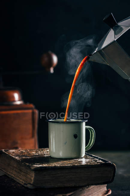 Poring hot coffee in enamel cup on old shabby books on dark background — Stock Photo