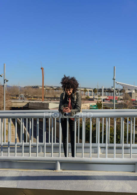 Frau mit Afrohaaren blickt auf ihr Handy, während sie auf einer Brücke in einer Stadt spaziert — Stockfoto
