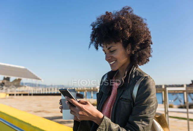 Черная женщина с афроволосами, прислонившаяся к ярко окрашенным стенам, глядя на свой смартфон и выпивая кофе — стоковое фото