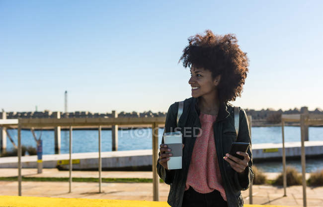 Black woman with afro hair leaning against brightly colored walls while looking at her smartphone and having a coffee — Stock Photo