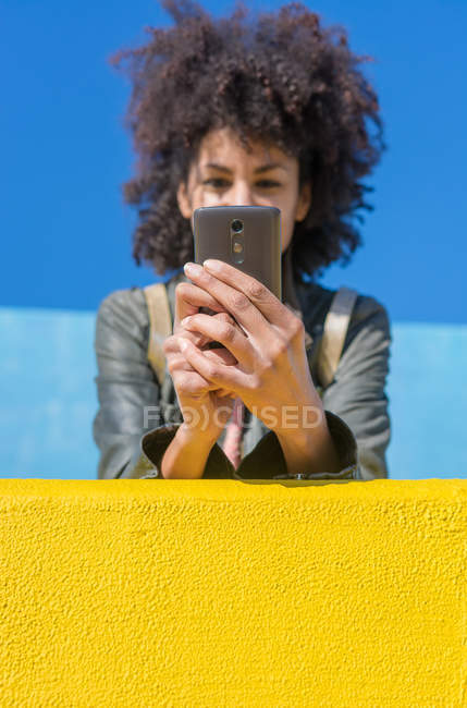 Donna nera con i capelli afro appoggiata alle pareti dai colori vivaci mentre guarda il suo smartphone e prende un caffè — Foto stock