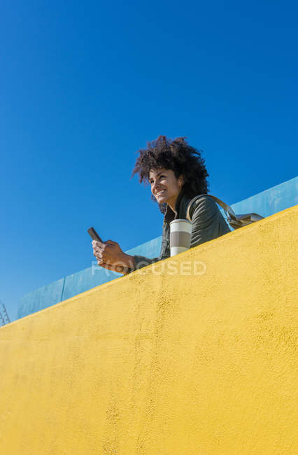 Femme noire aux cheveux afro s'appuyant sur des murs aux couleurs vives tout en regardant son smartphone et en prenant un café — Photo de stock
