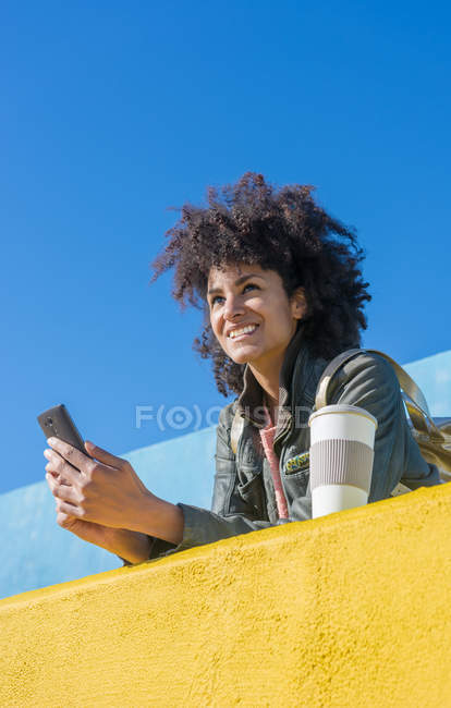 Schwarze Frau mit Afro-Haaren lehnt an bunten Wänden, während sie auf ihr Smartphone schaut und einen Kaffee trinkt — Stockfoto