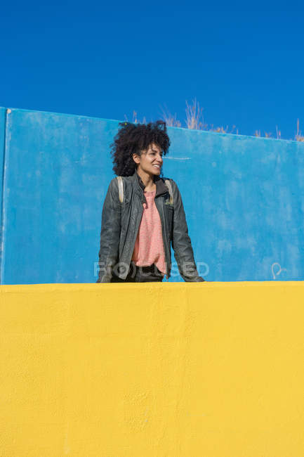 Mulher negra com cabelo afro encostado contra paredes coloridas enquanto olha para o smartphone e toma um café — Fotografia de Stock