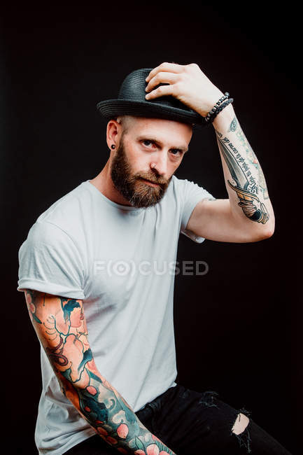 Cheerful bearded hipster in hat and t-shirt with tattoos on arms on black background — Stock Photo