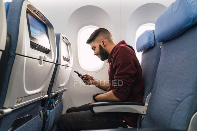 Vista lateral del guapo teléfono inteligente barbudo de navegación masculina mientras está sentado en un asiento cómodo dentro de aviones modernos - foto de stock