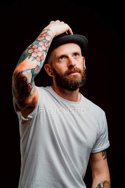 Cheerful bearded hipster in hat and t-shirt with tattoos on arms on black background — Stock Photo