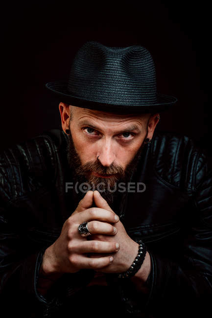 Cool hipster in hat and leather jacket looking at camera on black background — Stock Photo