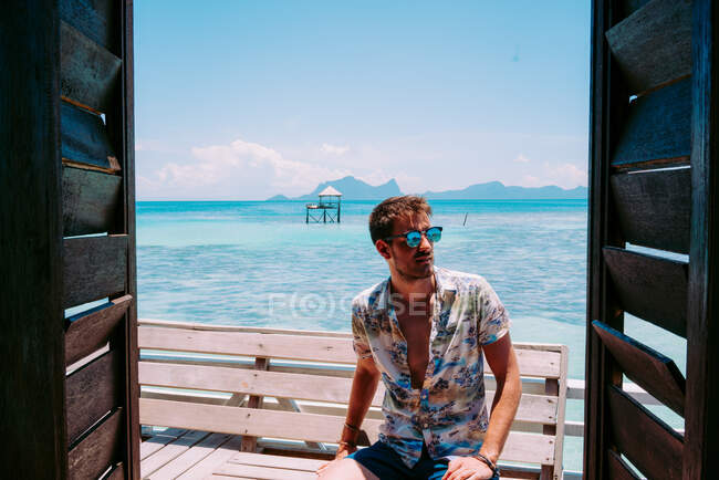 Junger Mann mit Sonnenbrille sitzt auf Sitz in der Nähe des blauen Meeres und schaut weg in Jamaica — Stockfoto