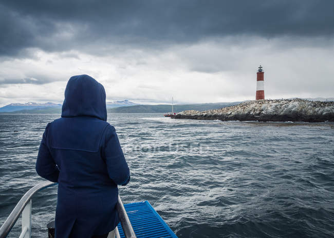 Vista posteriore di persona in felpa con cappuccio guardando il faro mentre in piedi sulla nave in mare tempestoso il giorno coperto — Foto stock