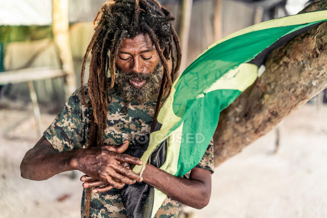 Varón barbudo afroamericano con rastas sosteniendo bandera de Jamaica cerca del árbol — Stock Photo