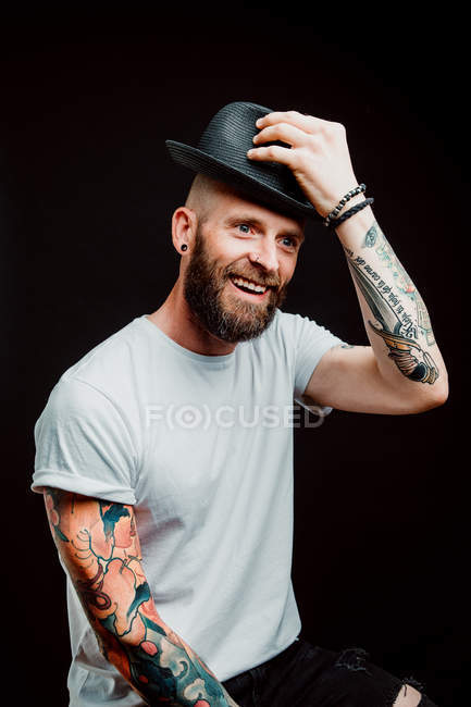 Alegre hipster barbudo em chapéu e t-shirt com tatuagens nos braços em fundo preto — Fotografia de Stock