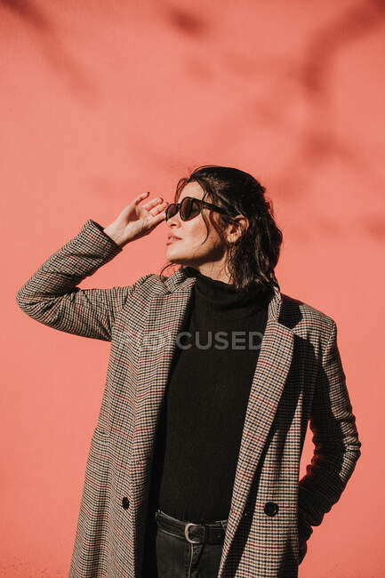 Beautiful trendy young woman in coat holding sunglasses and looking away on pink background — Stock Photo