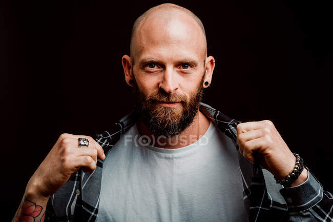 Hipster sin pelo barbudo en camisa con tatuajes en brazos sobre fondo negro - foto de stock