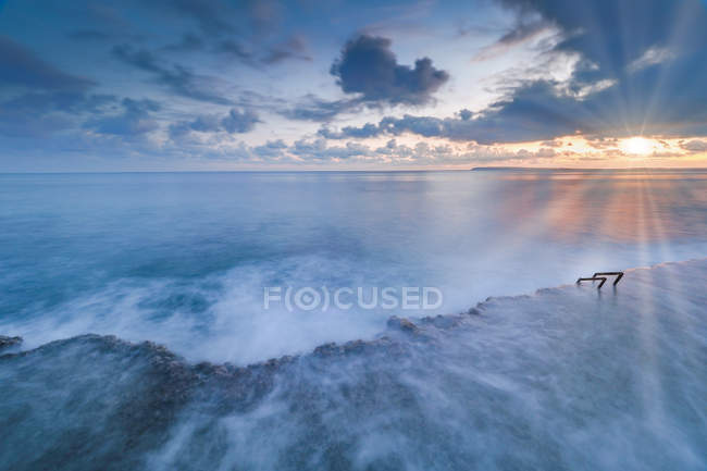 Luminoso sole che sorge sul cielo nuvoloso sul meraviglioso mare che ondeggia vicino alla costa rocciosa in natura — Foto stock