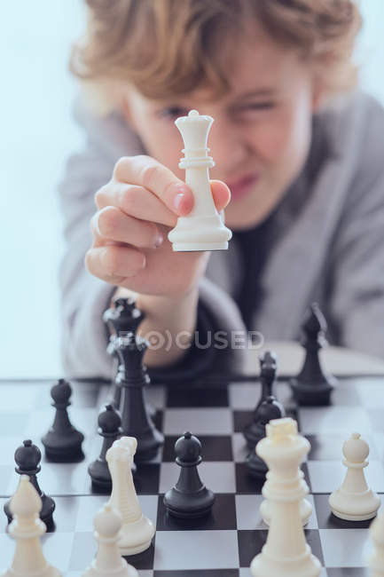 Criança engraçada segurando figura de xadrez branco perto do tabuleiro de xadrez no fundo turvo — Fotografia de Stock