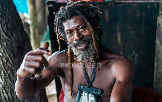 Afro-Américain mâle barbu avec dreadlocks et mains surélevées fumant cigare près des arbres — Photo de stock