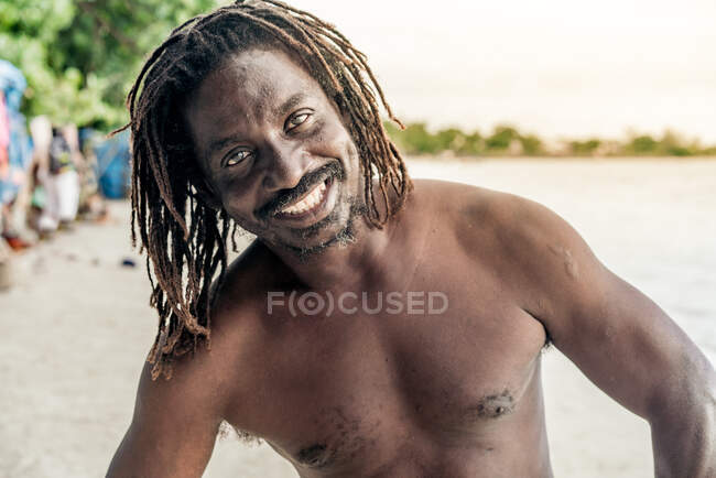 Веселый африканский мужчина без рубашки смотрит в камеру на размытом фоне на Ямайке — стоковое фото