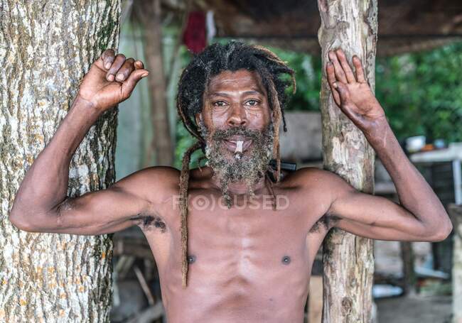 Hombre barbudo afroamericano con rastas y manos levantadas fumando cigarro cerca de los árboles - foto de stock