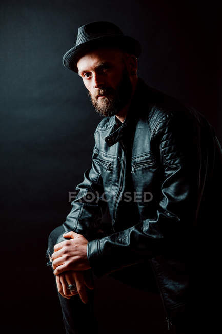 Cool hipster in hat and leather jacket looking at camera on black background — Stock Photo