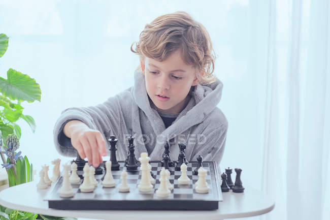 Kind hält weiße Figur auf Schachbrett am Tisch neben weißen Vorhängen — Stockfoto