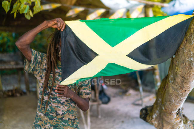 Homme barbu afro-américain avec dreadlocks tenant le drapeau jamaïcain près d'un arbre — Photo de stock