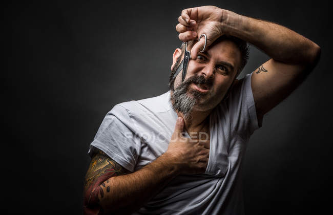 Adult man rubbing chest and looking at camera while cutting beard with scissors and standing on gray background — Stock Photo