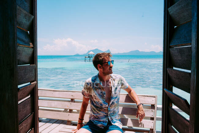 Молодой парень в солнцезащитных очках, сидящий на сиденье у синего моря и отводящий взгляд в ямайке — стоковое фото