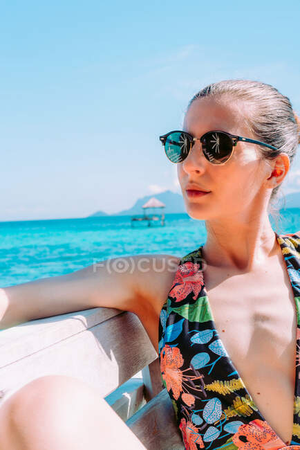 Дама в купальнику і сонячні окуляри сидять на сидінні біля блакитного моря в Ямайці. — стокове фото
