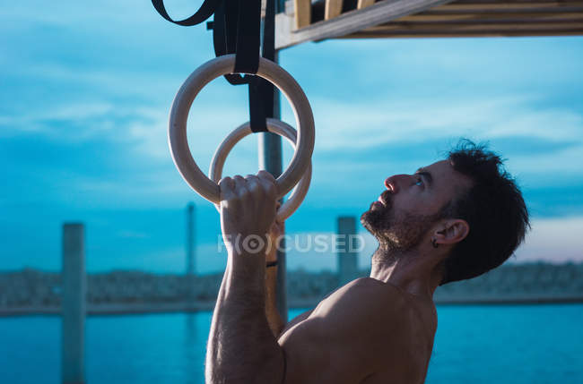 Athletic man balancing on gymnastic rings on embankment in city — Stock Photo