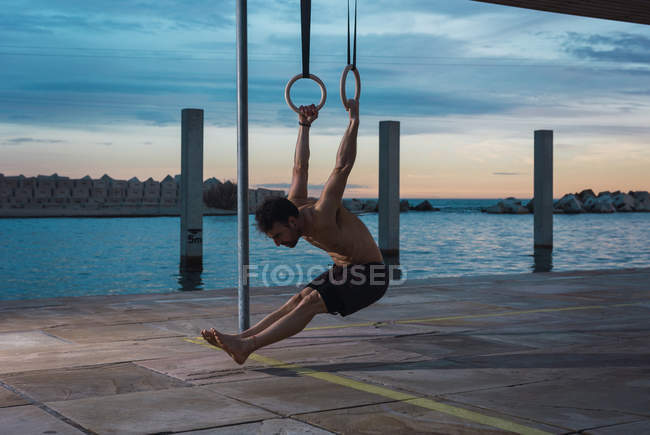 Атлетик балансирует на гимнастических кольцах на набережной в вечернем городе — стоковое фото
