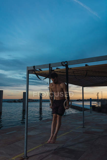 Athletic man balancing on gymnastic rings on embankment in evening city — Stock Photo