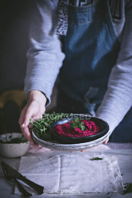 Человеческие руки, подающие домашнюю свеклу хумус на тарелке с травами — стоковое фото