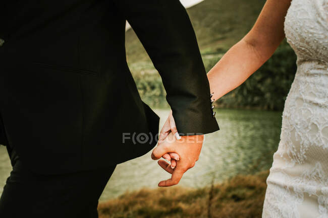 Hombre y mujer sin rostro usando prendas de boda y tomados de la mano mientras caminan por la costa - foto de stock