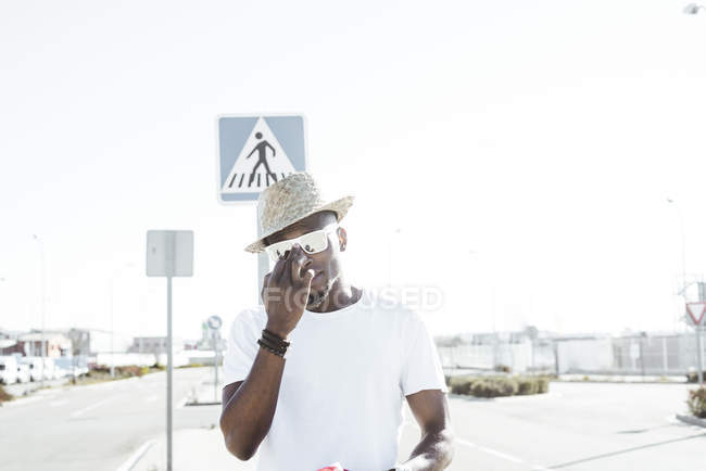 Hombre negro de moda en sombrero y gafas de sol de pie en la luz brillante en la calle mirando a la cámara - foto de stock