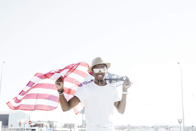 Content stylish black man in sunglasses and hat holding American flag smiling at camera in sunlight — Stock Photo