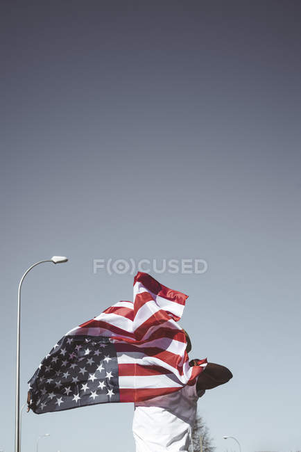 From below of man in white t-shirt standing with waving American flag under blue sky on street — Stock Photo
