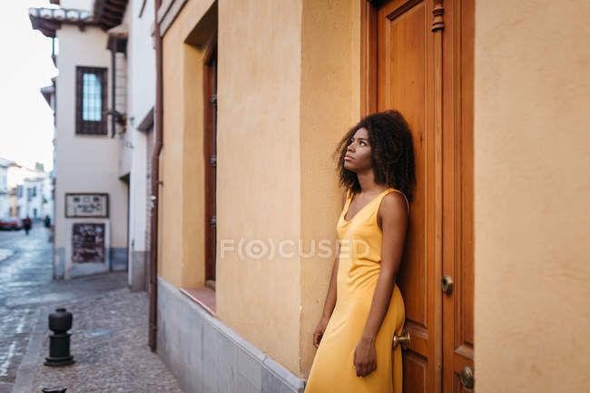 Pensive black woman in yellow dress leaning on door on street — Stock Photo
