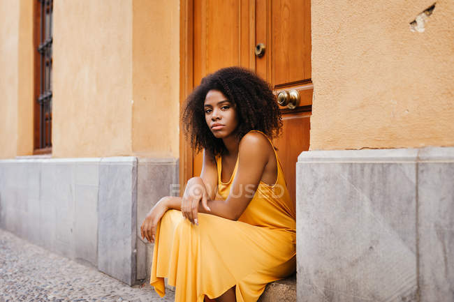 Gorgeous black woman in dress sitting on porch on street and looking at camera — Stock Photo