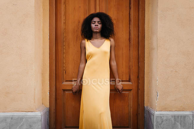 Femme noire rêveuse en robe jaune penchée sur la porte dans la rue — Photo de stock