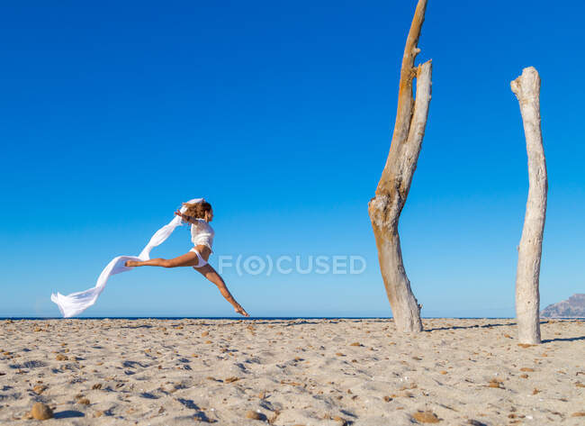 Vista lateral de la atractiva mujer saltando con pareo en la playa de arena posada día soleado sin nubes - foto de stock