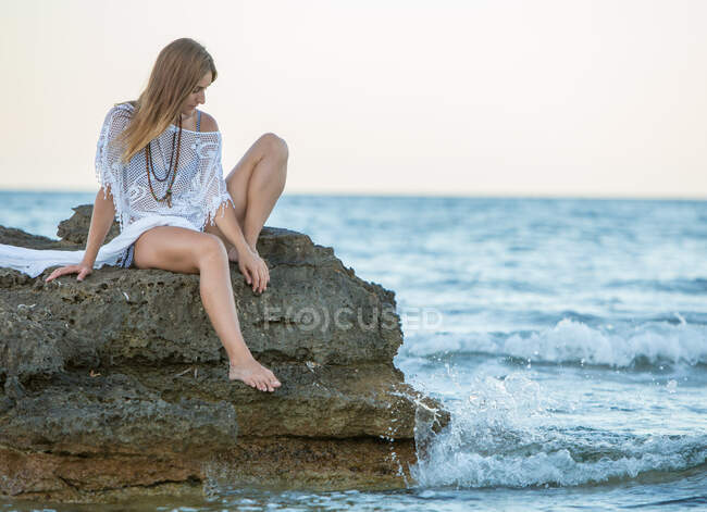 Mujer joven soñando despierta en ropa de playa sentada en piedra a la orilla del mar y mirando a la cámara - foto de stock