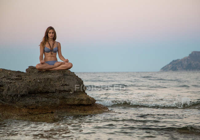 Mujer joven soñando despierta en ropa de playa sentada en postura de meditación sobre piedra en la orilla del mar - foto de stock