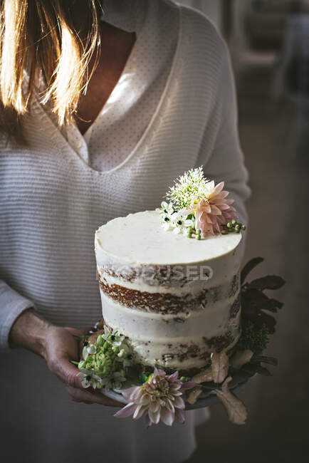 Side view of crop happy lady holding dish with tasty cake decorated by chrysanthemum bud and dry leaves in room — Stock Photo