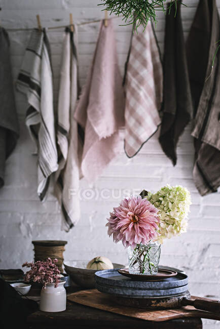 Wooden table with bunch of fresh pink chrysanthemums and white hydrangea in vase between frying pan and kitchenware near dish cloths hanging on twist with pins — Stock Photo