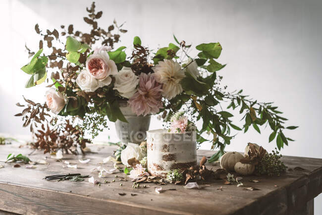 Dish with tasty cake decorated bloom bud on wooden table with bunch of chrysanthemums, roses and plant twigs in vase between dry leaves on grey background — Stock Photo