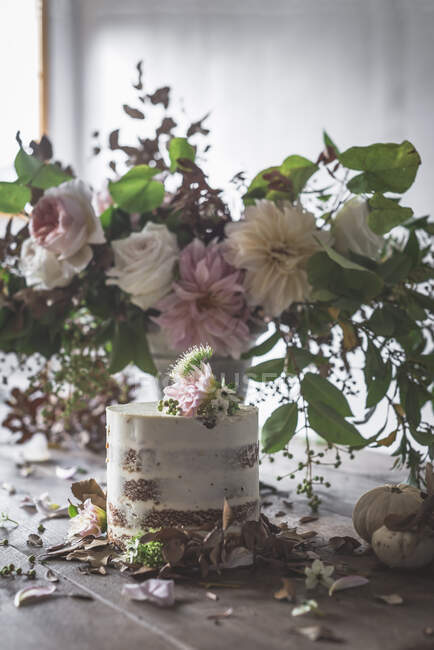 Plat avec gâteau savoureux bourgeon fleuri décoré sur table en bois avec bouquet de chrysanthèmes, roses et brindilles de plantes dans un vase entre les feuilles sèches sur fond gris — Photo de stock